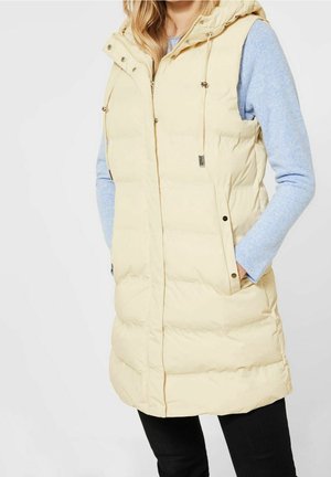 Kobieta ubrana w długą beżową kamizelkę puchową z kapturem na jasnoniebieskim swetrze z długim rękawem, ręce w kieszeniach, na jednolitym tle.