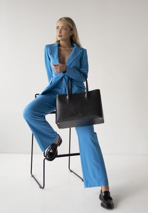 Borsa a spalla nera in pelle con texture liscia, due manici rotondi e dettagli del logo. Il modello indossa un completo azzurro chiaro e mocassini neri, seduto.