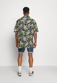 Camisa de manga corta con estampado floral oscuro que presenta patrones tropicales en rosa y verde, combinada con pantalones cortos de denim enrollados y zapatillas deportivas.