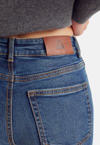 Jeans de mezclilla azul con un ajuste de cintura alta, con un parche de cuero marrón en la parte posterior que presenta un emblema de pulpo y acentos de costura naranja.