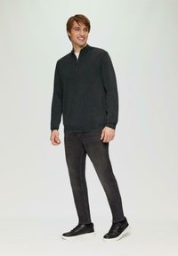 Donkergrijze ritstrui van textuurstof, gecombineerd met slim-fit zwarte jeans en zwarte sneakers met witte zolen, staand tegen een lichte achtergrond.
