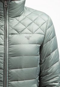 Quiltet grøn jakke med høj krave, der har en frontlynlås og 'GANT' logoet broderet på brystet. Glat, blank overflade.
