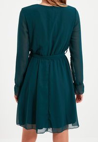 Robe teal avec de longues manches transparentes, une taille cintrée et une jupe évasée. Tissu lisse avec un détail de ceinture à la taille.