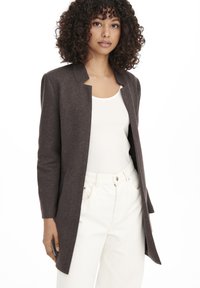 Blazer lungo, di colore marrone scuro, su misura, con collo a caduta, aperture frontali e tessuto testurizzato. Indossato sopra una maglietta bianca e pantaloni chiari.