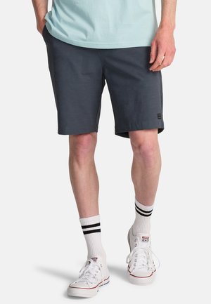 Mężczyzna w ciemnoszarych szortach, białych skarpetkach za kostkę z czarnymi paskami oraz białych trampkach Converse, stojący z rękami w kieszeniach.