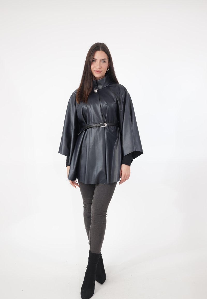Schwarzer Cape-Mantel aus Kunstleder mit hohem Kragen, Gürtel in der Taille und weiten Ärmeln. Kombiniert mit grauen, eng anliegenden Jeans und schwarzen Stiefeletten.