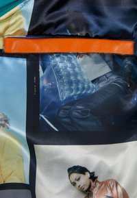 Saténová látka s barevným koláží fotografických tisků. Silný oranžový kožený popruh je výrazně umístěn nahoře.