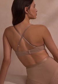 Donna vista da dietro che indossa un reggiseno beige con spalline incrociate e regolabili e biancheria intima a vita alta abbinata su uno sfondo neutro.