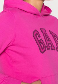 Sudadera rosa de algodón, que presenta un gran logo de "GAP" en costura morada oscura. Incluye un bolsillo frontal y capucha con cordón.
