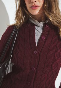 Burgundijas krāsu adīta kardigāna jakas ar V-veida kakla izgriezumu un purpura pogām, pārklāta virs pelēka augstākās kakla izgriezuma krekla, saskaņota ar melnu plecu somu.