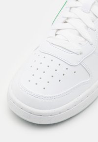 White leather sneaker with textured upper, rounded toe, perforated detailing, and green accent near the laces. Rubber sole with visible stitching.