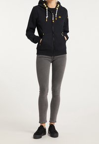 Felpa nera con zip e tasca frontale, con dettagli gialli sui lacci e sul logo. Abbinata a jeans skinny grigi e sneakers nere.