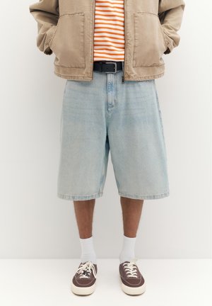 Personne portant un short en jean bleu clair, des baskets marron avec des lacets blancs, des chaussettes blanches, une veste beige et une chemise rayée orange.