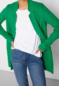 Cárdigan verde de punto con cuello, abertura delantera y mangas largas. Llevado sobre una camisa blanca y combinado con jeans azules. Diseño sencillo.