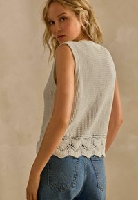 Hellgraues gestricktes Tanktop mit gewelltem Saum und strukturiertem Muster. Das Modell trägt eine blaue Jeans und ist von hinten zu sehen.