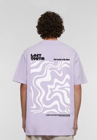 Lilla t-skjorte med korte ermer som har et bølgete hvitt grafisk design og svart tekst på baksiden. Myk bomullsstoff med en avslappet passform.