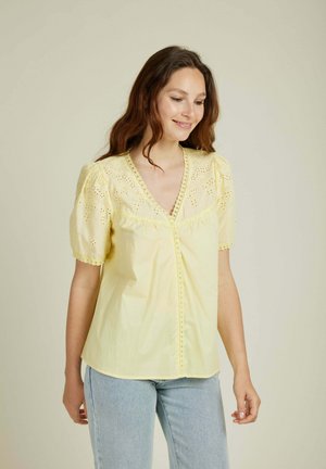 Blouse jaune avec un décolleté plongeant et des manches courtes bouffantes. Présente des broderies à jour et des boutons décoratifs sur le devant, fabriquée en tissu léger.