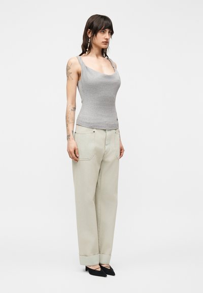 Γυναίκα με σκούρα μαλλιά μέχρι τους ώμους που φοράει γκρι εφαρμοστό τοπ χωρίς μανίκια, ανοιχτό μπεζ παντελόνι με φαρδιά γραμμή, μαύρες μυτερές γόβες και μακριά σκουλαρίκια.