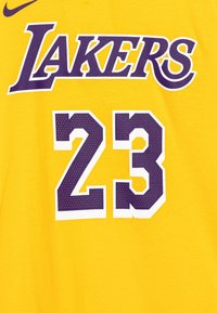 Keltainen puuvillapaita, jossa on "LAKERS" purppuran värillä suuremman, tekstuurilla varustetun valkoisen ääriviivan "23" päällä purppuran värillä, urheilullisella designilla.