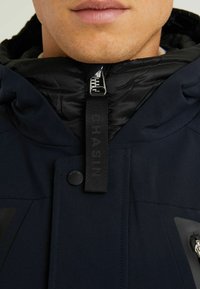 Granatowa kurtka z odpinaną, czarną, pikowaną warstwą wewnętrzną. Posiada zamek błyskawiczny, oznaczoną zawieszkę oraz zatrzaski. Miękka w dotyku.