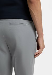 Jasnoszare spodnie o teksturowanym wykończeniu, z tylną kieszenią i subtelną metką z logo. Gładka tkanina i dopasowany krój widoczne.