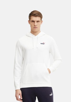 Giovane uomo che indossa una felpa con cappuccio bianca Puma con logo sul petto e pantaloni della tuta neri Puma, in piedi davanti a uno sfondo chiaro e semplice.