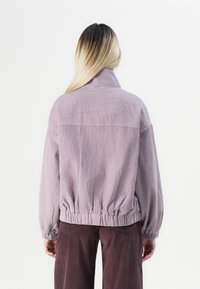 Persoon met lang ombre haar, gekleed in een lichtpaarse corduroy jas en een donkerpaarse corduroy broek, gezien vanachter tegen een witte achtergrond.