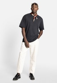 Bărbat stând cu o mână în buzunar, purtând un tricou negru, pantaloni albi, pantofi negri cu șireturi, o eșarfă roșie cu model la gât și ochelari de soare aurii pe cap.