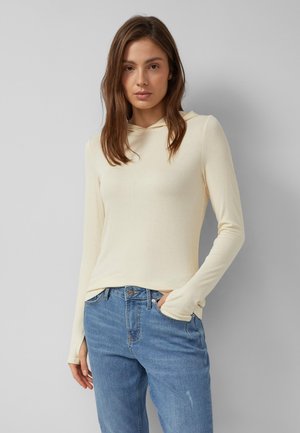 Vrouw staand tegen een muur, gekleed in een crèmekleurige, lange mouwen hoodie en blauwe spijkerbroek, met één hand in haar zak.