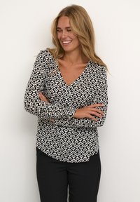 Blusa a maniche lunghe bianca e nera con scollatura a V, caratterizzata da motivi geometrici e orlo curvo. Abbinata a pantaloni neri.