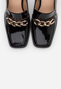 Mocassins en cuir verni noir avec des orteils carrés, présentant un accent en chaîne dorée sur le dessus et des semelles extérieures noires texturées.