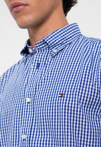 Niebieska i biała koszula w kratkę w stylu gingham, zapinana na guziki, z kołnierzykiem, z małym logo w czerwonym, białym i niebieskim kolorze na piersi.