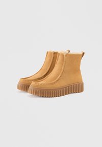 Une paire de bottes cheville en daim beige avec des semelles en caoutchouc ribbées épaisses et une doublure en polaire crème visible sur les bords supérieurs.