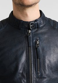 Námořnická modrá kožená bunda s vysokým límcem, výraznými zipy a texturovanými akcenty na ramenou. Hladká textura, elegantní design.
