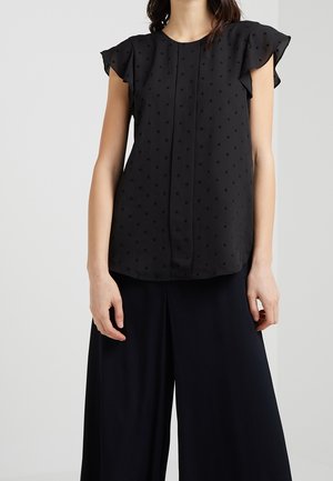 Vrouw die een zwarte mouwloze blouse met kleine sterpatronen en zwarte wijde broek draagt tegen een effen lichte achtergrond.