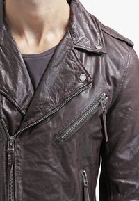Giacca da motociclista in pelle marrone con una finitura testurizzata. Presenta tasche con zip ed epaulette. Chiusura frontale asimmetrica con zip e bottoni a pressione.