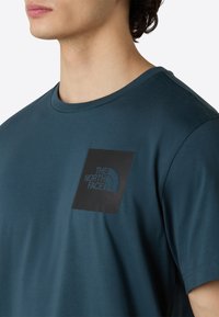 Teal bomulds t-shirt med rund hals og korte ærmer. Har et sort firkantet plade med den hvide "The North Face" logo. Blød tekstur.