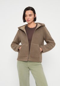 Καφέ φλις μπουφάν με κουκούλα, υφή με ανάγλυφη επιφάνεια, κλείσιμο με φερμουάρ και δύο εμπρόσθιες τσέπες. Φοριέται πάνω από ένα σκούρο πουκάμισο και ανοιχτόχρωμο παντελόνι.