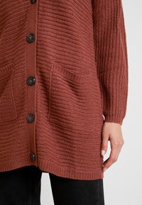 Strickjacke in Rostfarbe mit großen schwarzen Knöpfen, zwei Vorderentaschen und gerippten Bündchen, die ein strukturiertes Stoffdesign hervorheben.