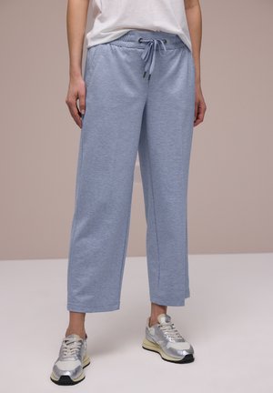 Kvinde iført lyseblå vidde croppede sweatpants med snoretræk i taljen og hvide og sølvfarvede sneakers, stående mod en ensfarvet baggrund.