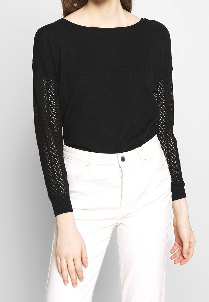 Femme portant un haut noir à manches longues avec des manches transparentes à motifs et un pantalon blanc taille haute sur un fond uni.