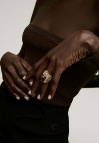 Anillo texturizado en tono dorado con diseño de hoja, llevado en una mano de piel oscura con uñas cortas y bien cuidadas; sobre un top marrón.