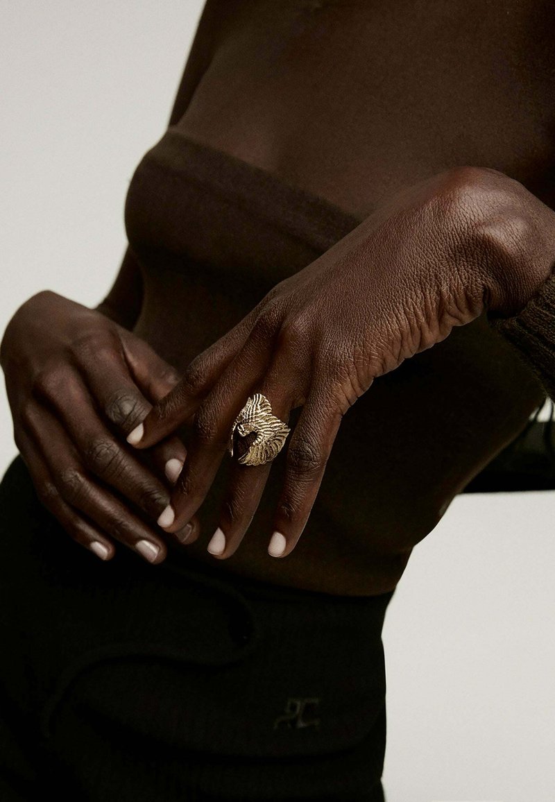 Anillo texturizado en tono dorado con diseño de hoja, llevado en una mano de piel oscura con uñas cortas y bien cuidadas; sobre un top marrón.