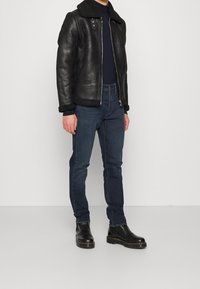 Musta nahkatakki, jossa on shearling-kaulus, yhdistetty tummansinihin farkkuihin ja mustiin nahkabootseihin. Takissa on vetoketjusuljenta ja nepparit.