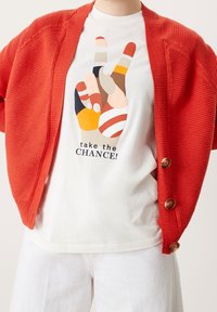 Röd ribbad kofta med träknappar över en vit t-shirt med en flerfärgad handgrafik och texten "ta CHANSEN!" i svart.