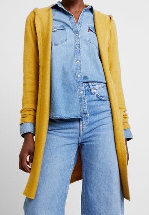 Henkilö pukeutunut vaaleansiniseen farkkupaitaan ja farkkuihin sekä sinappikeltaiseen pitkään neuletakkiin, seisoo yksiväristä taustaa vasten.