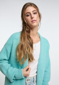 Turkos stickad cardigan med öppen framsida, ribbade manschetter och mjuk textur; kombinerad med en vit t-shirt och ljusblå jeans.