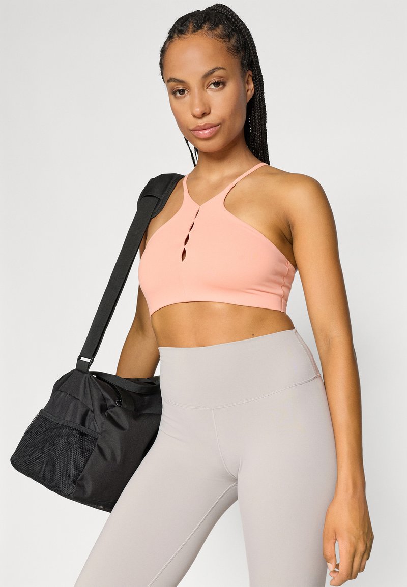 Helles rosa Sport-BH mit Schlüssel-Loch-Detail, kombiniert mit hochgeschnittenen, hellgrauen Leggings; schwarze Sporttasche über die Schulter gehängt.