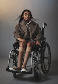 Jeune femme aux longs cheveux et manteau marron assise dans un fauteuil roulant devant un fond gris, regardant devant elle, les mains posées sur ses genoux.
