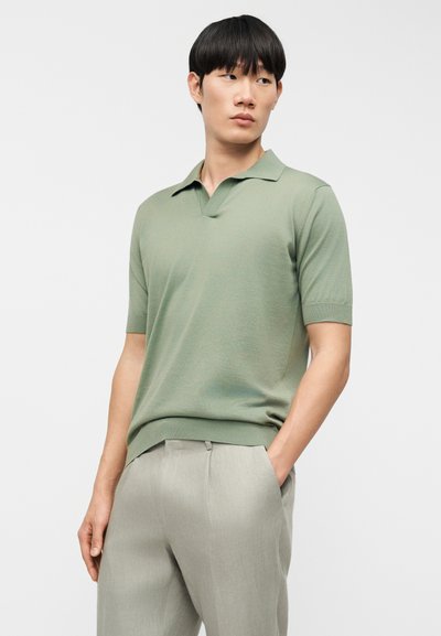 Πόλο μπλούζα με κοντό μανίκι από πλεκτό υλικό ανοιχτού πράσινου, με V-λαιμόκοψη και ριμπ μανσέτες, συνδυασμένη με ανοιχτό γκρι παντελόνι καστοριού.